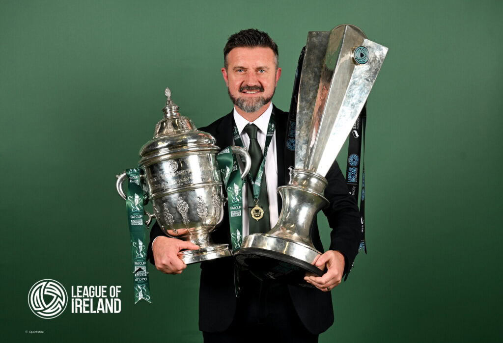 Champions League trophy held by smiling man in suit with League of Ireland logo in background.