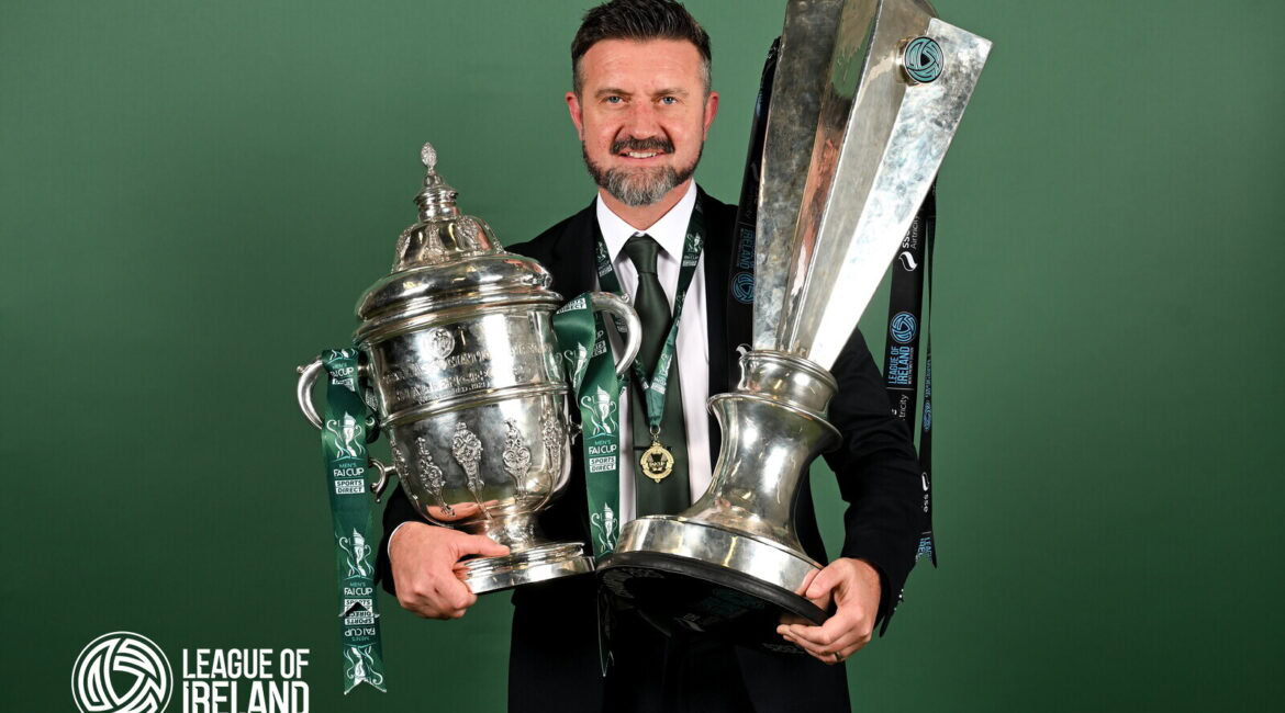 Champions League trophy held by smiling man in suit with League of Ireland logo in background.