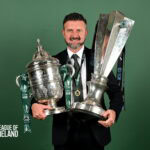 Champions League trophy held by smiling man in suit with League of Ireland logo in background.