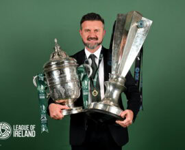 Champions League trophy held by smiling man in suit with League of Ireland logo in background.