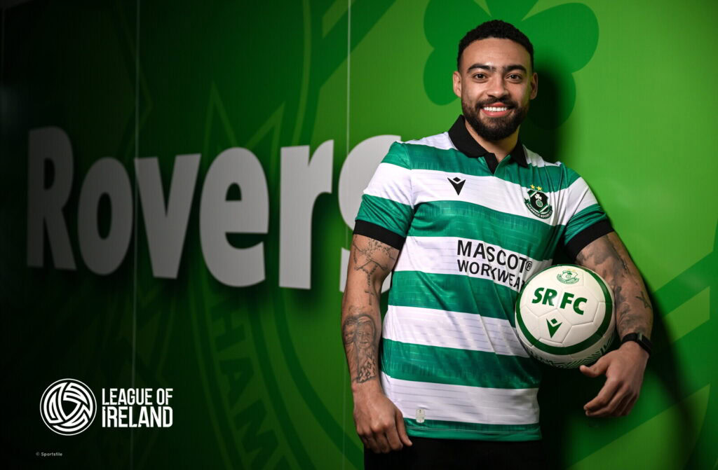 Bright smiling footballer in Shamrock Rovers jersey holding a soccer ball, standing against team branding and League of Ireland logo.