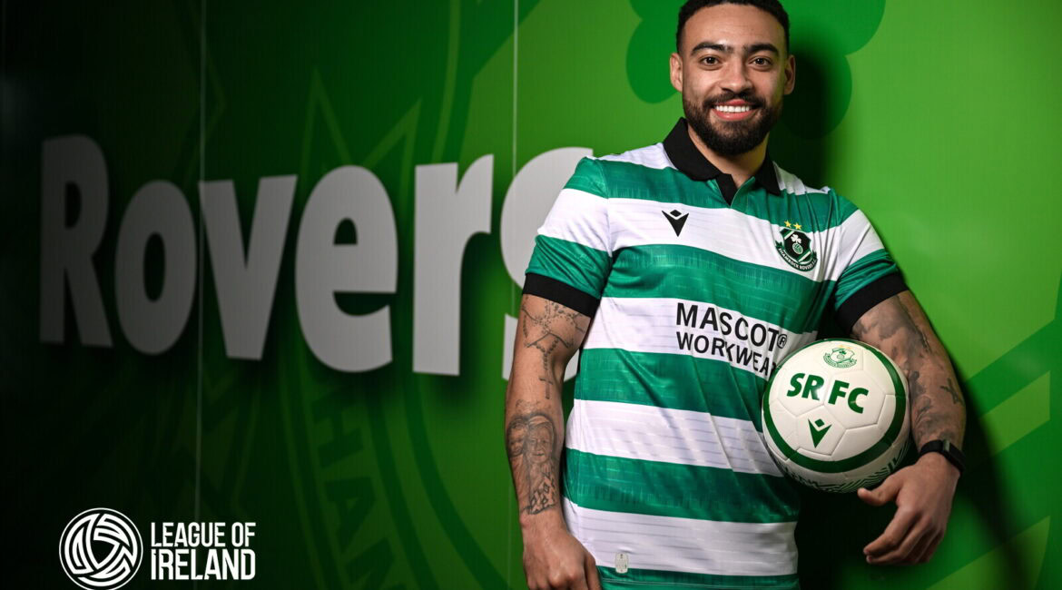 Bright smiling footballer in Shamrock Rovers jersey holding a soccer ball, standing against team branding and League of Ireland logo.
