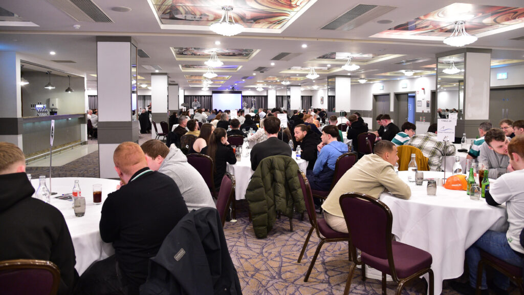 Brightly lit conference room filled with young men and women seated at round tables during a formal event.