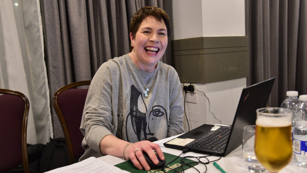 Smiling woman working on a laptop at a table with drinks and papers, promoting Shamrock Rovers football club.