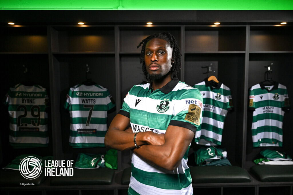 Shamrock Rovers player in locker room with team jerseys behind him.