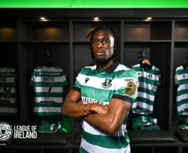 Shamrock Rovers player in locker room with team jerseys behind him.