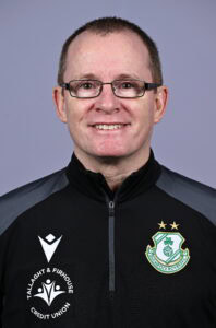 Shamrock Rovers coach smiling in team jacket with club crest, close-up portrait.