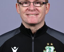 Shamrock Rovers coach smiling in team jacket with club crest, close-up portrait.