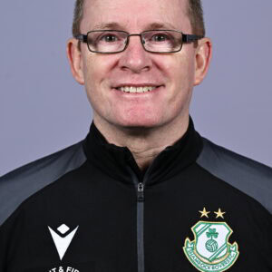 Shamrock Rovers coach smiling in team jacket with club crest, close-up portrait.