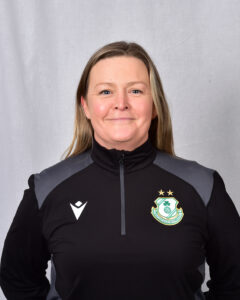 Female Shamrock Rovers coach in team jacket, smiling, standing against grey background.