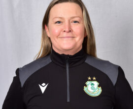 Female Shamrock Rovers coach in team jacket, smiling, standing against grey background.