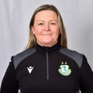 Female Shamrock Rovers coach in team jacket, smiling, standing against grey background.