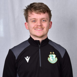 Shamrock Rovers football player smiling in team kit with club crest, standing against grey backgroun.