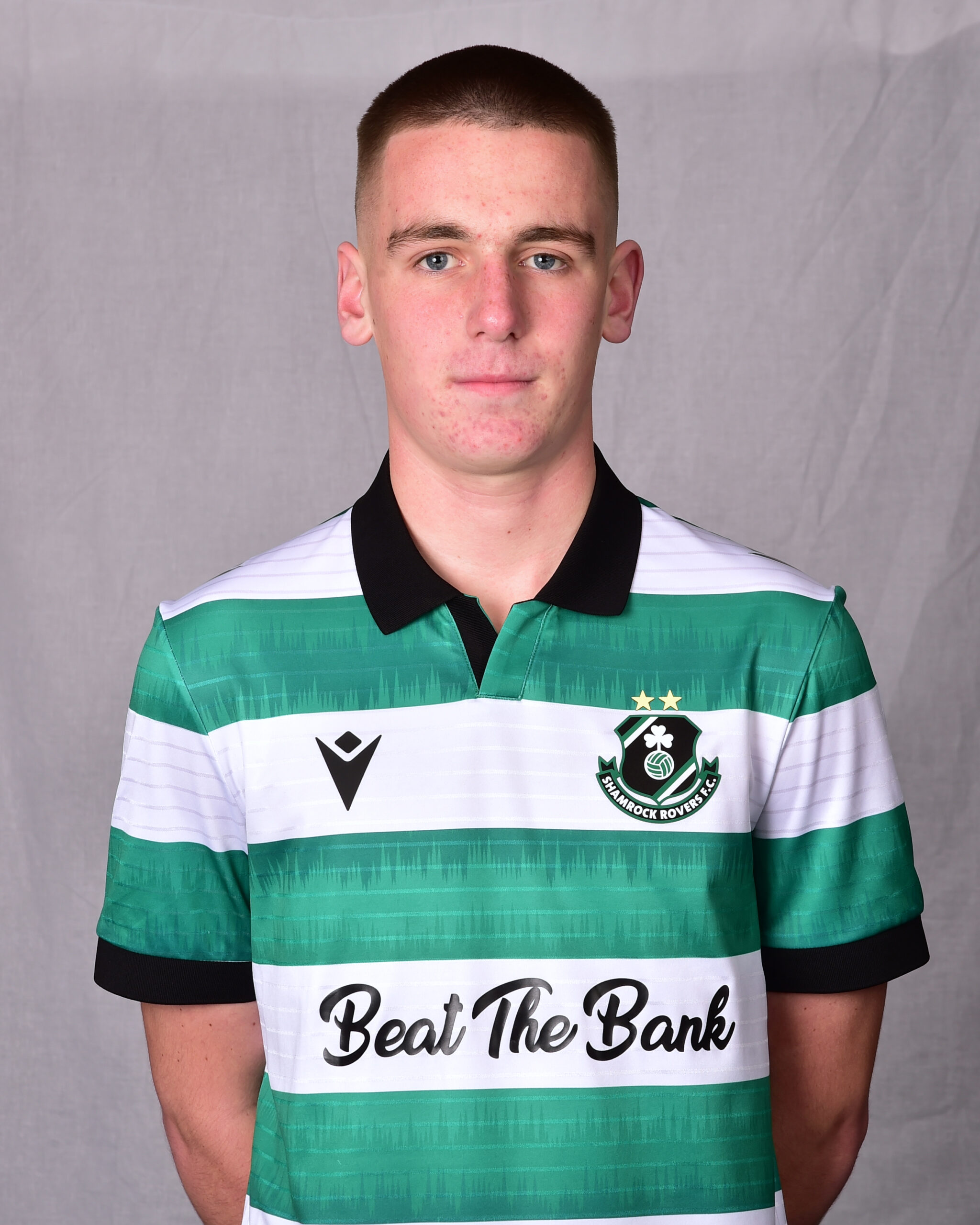 Young Shamrock Rovers player in green and white striped jersey, standing on grey background.