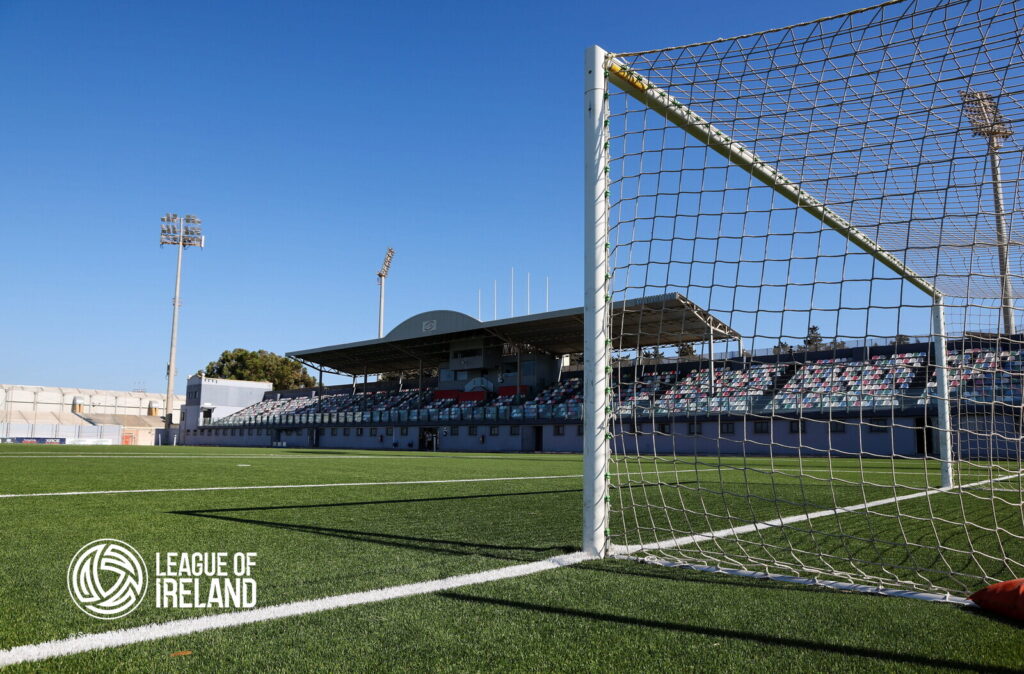 Empty football stadium with goalpost and League of Ireland logo.