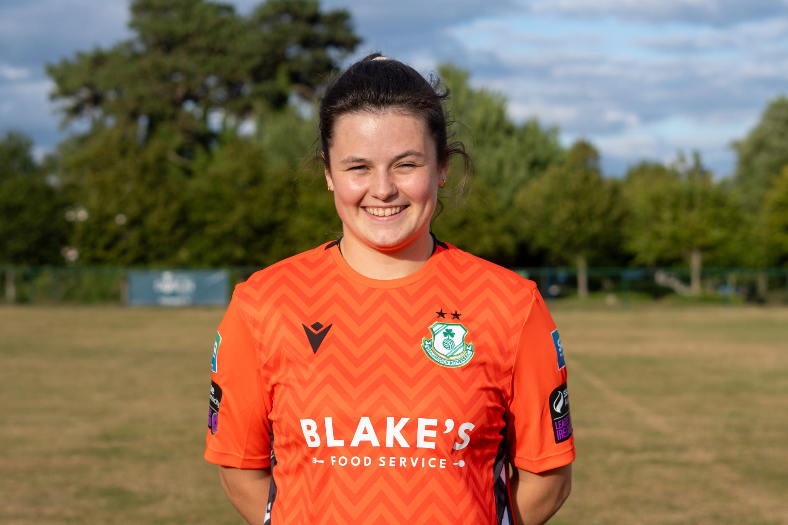 Shamrock Rovers female player in orange jersey smiling outdoors.